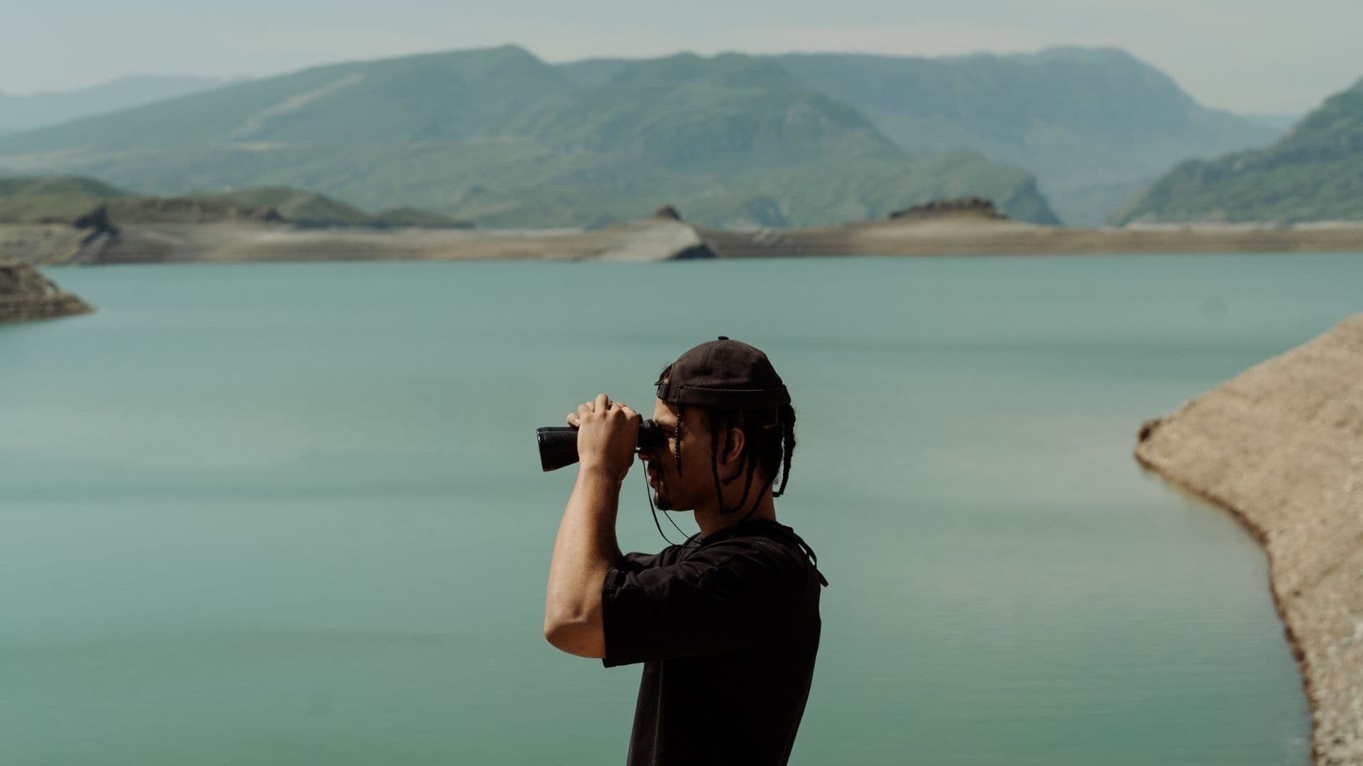 Un observateur en t-shirt noir scrute un lac turquoise et des montagnes aux jumelles, en plein jour