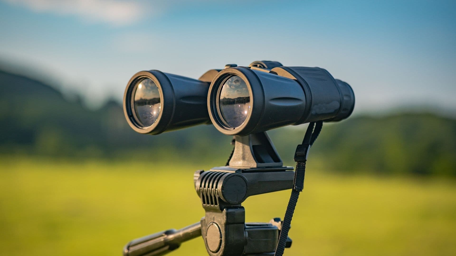 Close-up of a pair of binoculars on a tripod, coated lenses facing a green landscape